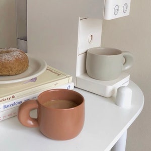 Peut inclure: Nature morte minimaliste avec une cafeti&egrave;re, deux tasses en c&eacute;ramique, une assiette avec un bagel et des livres. Les tasses sont dans des tons bruns et verts sourds. La sc&egrave;ne se d&eacute;roule sur une table blanche, cr&eacute;ant une esth&eacute;tique neutre.