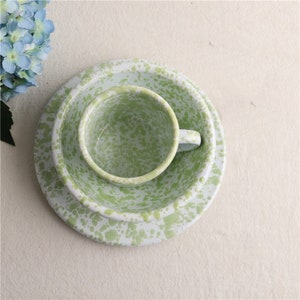 Peut inclure: Un service de vaisselle en émail tacheté vert et blanc, comprenant une assiette, un bol et une tasse. L'ensemble a une esthétique vintage et est disposé sur une surface neutre. La tasse est à l'intérieur du bol, qui est sur l'assiette.