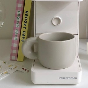 Peut inclure: Une tasse en c&eacute;ramique gris clair avec une forme arrondie et une grande anse circulaire. La tasse est pos&eacute;e sur une machine &agrave; expresso blanche. Deux livres avec le texte "GROOVY DAYS" sont visibles.