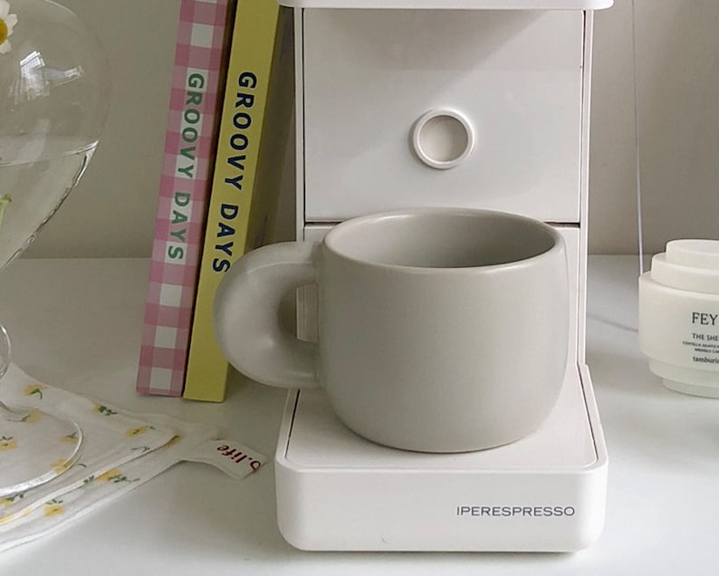Peut inclure: Une tasse en c&eacute;ramique gris clair avec une forme arrondie et une grande anse circulaire. La tasse est pos&eacute;e sur une machine &agrave; expresso blanche. Deux livres avec le texte "GROOVY DAYS" sont visibles.
