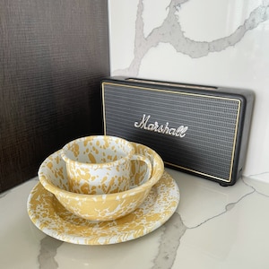 Peut inclure: Un service de vaisselle en émail moucheté jaune et blanc, comprenant une tasse, un bol et une assiette. Un haut-parleur Marshall noir avec des bordures dorées est en arrière-plan. L'ensemble est sur un comptoir blanc.