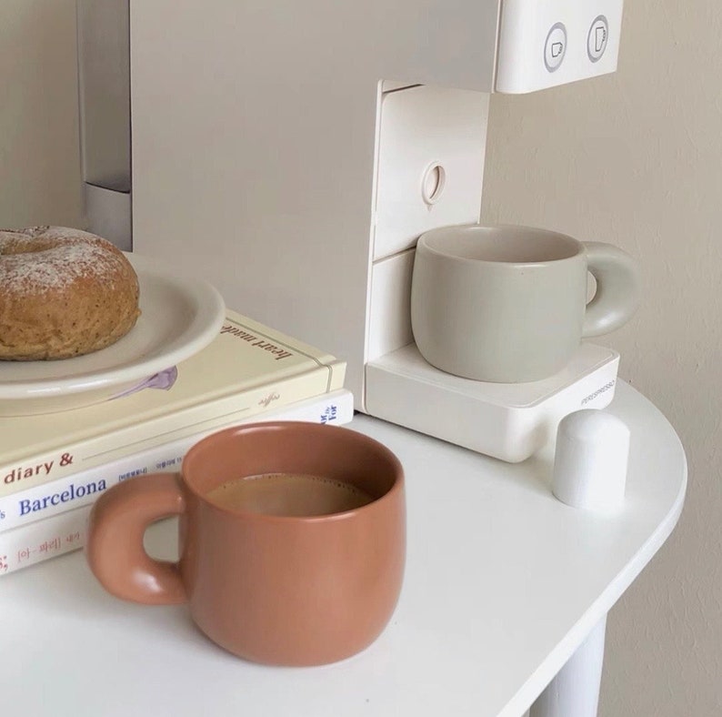 Peut inclure: Nature morte minimaliste avec une cafeti&egrave;re, deux tasses en c&eacute;ramique, une assiette avec un bagel et des livres. Les tasses sont dans des tons bruns et verts sourds. La sc&egrave;ne se d&eacute;roule sur une table blanche, cr&eacute;ant une esth&eacute;tique neutre.