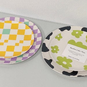 Peut inclure: Deux assiettes décoratives. L'une présente un motif à carreaux jaunes et blancs sur un fond à carreaux violets. L'autre a un fond blanc avec des fleurs vertes et des taches noires ressemblant à des vaches. Une étiquette est visible.