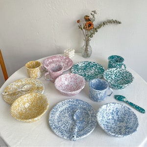 May include: A collection of colorful enamelware dishes, including plates, bowls, and mugs, with a speckled pattern. The set features various colors like yellow, pink, green, and blue. A spoon is included with some of the sets. The dishes are arranged on a white surface.