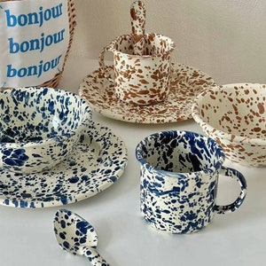 May include: A selection of speckled ceramic tableware in blue and brown. The set includes bowls, plates, a mug, and a spoon. The pieces have a white base with dark blue or brown splatters. A woven basket with the text "bonjour bonjour bonjour" is in the background.