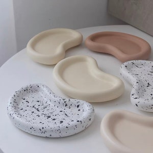 Peut inclure: Plusieurs plateaux en céramique en forme de rein, dans des tons crème, beige et blanc moucheté de noir. Les formes lisses et arrondies suggèrent un design moderne et minimaliste, idéal pour un usage décoratif ou fonctionnel.