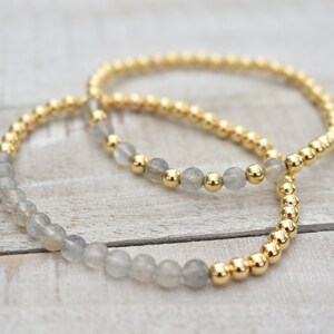 May include: Two bracelets, one with grey beads and one with gold beads, are layered on a light wood surface.
