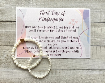May include: Two white pearl bracelets with gold heart charms and a card that reads "First Day of Kindergarten" with a poem about wearing the bracelets on the first day of school.