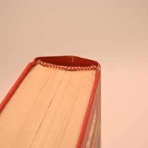 May include: Close-up of a book's corner, showcasing the red cover and pages. The binding is reinforced with a red and white patterned thread. The book's pages are a light beige color.