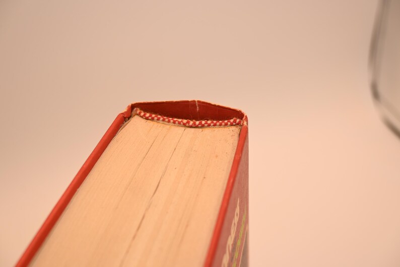May include: Close-up of a book's corner, showcasing the red cover and pages. The binding is reinforced with a red and white patterned thread. The book's pages are a light beige color.