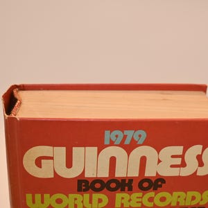 May include: A vintage 1979 Guinness Book of World Records. The book has a red cover with white and yellow lettering. The book's pages are a light tan color. The book is in good condition, with some wear and tear on the edges.