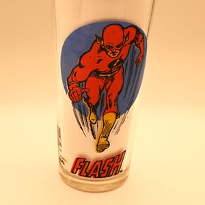 May include: A clear glass featuring a graphic of the Flash, a superhero in a red suit, running. The Flash is set against a blue oval background. The word "FLASH" is printed in red at the bottom.