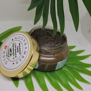 May include: A jar of dark brown skincare product with a gold lid, labeled "NOOR & AMBER Skin&Hair CARE All Natural Ingredients." The jar is open, revealing the product. Green leaves are in the background.
