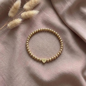 Könnte beinhalten: Goldenes Perlenarmband mit herzförmigem Anhänger. Der Anhänger trägt den Buchstaben "A". Das Armband wird auf einem rosa Stoff präsentiert, mit getrockneten Blumen im Hintergrund. Ein zartes Schmuckstück.