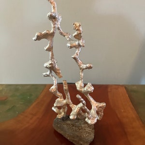 May include: A silver aluminum sculpture resembling a tree with a rough, gray base. The sculpture has a unique, organic shape and a textured surface.