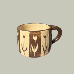 May include: A ceramic mug featuring a repeating pattern of dark brown vertical stripes alternating with cream-colored panels. Each cream panel displays a hand-painted dark brown tulip illustration. The mug has a thick, curved handle on the right side.