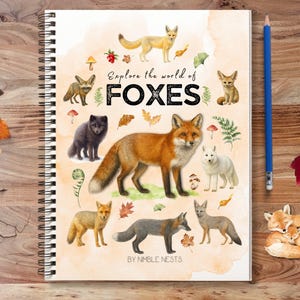 May include: A spiral-bound notebook featuring the text "Explore the world of FOXES" and illustrations of different fox species. A blue pencil and autumn leaves are placed next to the notebook on a wooden surface. The notebook has a watercolour design with foxes, mushrooms, and foliage.