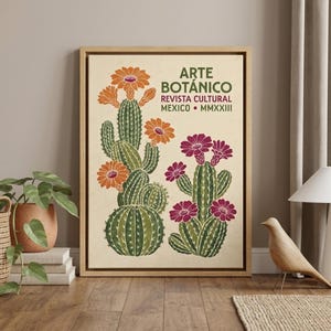 Op de afbeelding: Een ingelijste wandkunstprint met twee groene cactussen met oranje en magenta bloemen op een crèmekleurige achtergrond. De tekst luidt ARTE BOTÁNICO REVISTA CULTURAL MEXICO MMXXIII in donkergroen en rood. De print zit in een lichthouten lijst.