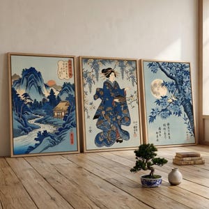 Op de afbeelding: Drie ingelijste Japanse houtsneden. De prenten tonen natuurtoneel, waaronder een berglandschap, een vrouw in kimono en een boom in maanlicht. De prenten zijn in blauw- en beigetinten, met houten frames.