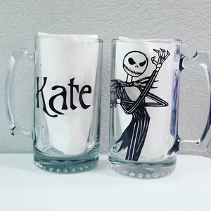 May include: Two clear glass beer mugs. One mug has the name "Kate" in black script. The other mug features a black and white illustration of Jack Skellington from The Nightmare Before Christmas.