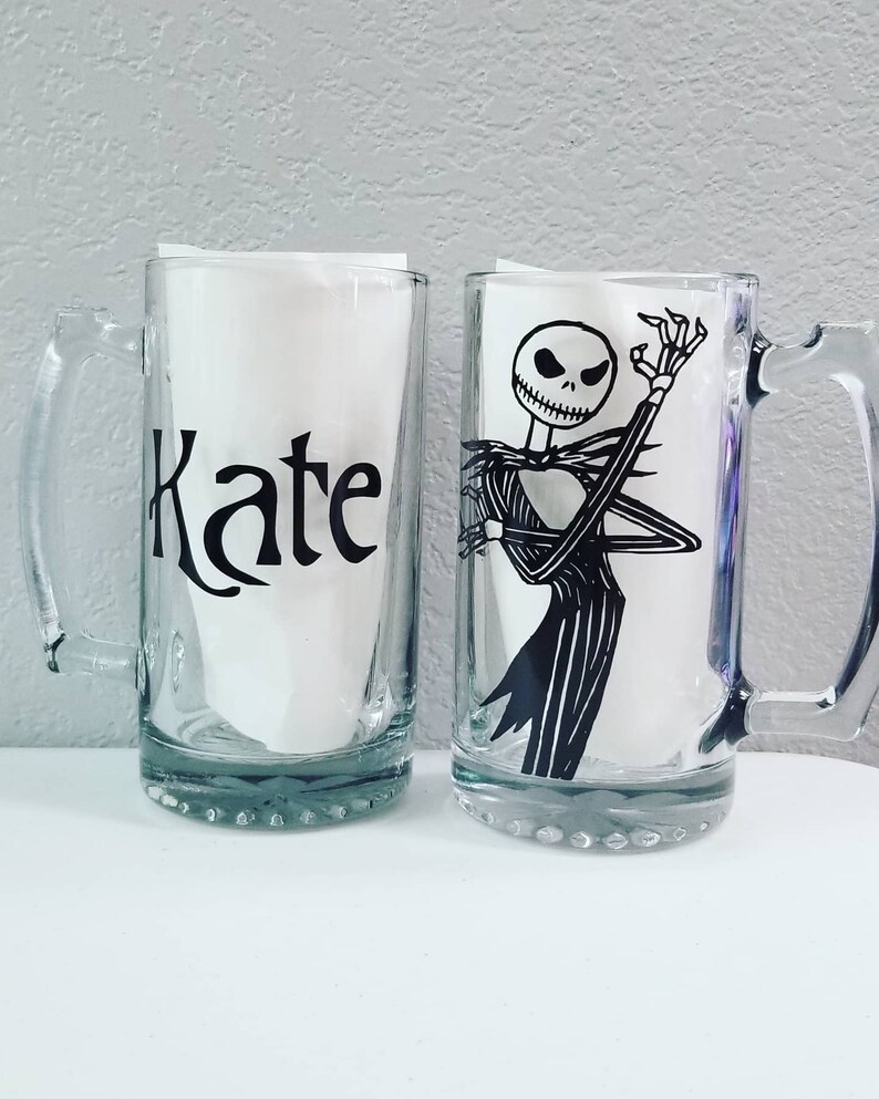 May include: Two clear glass beer mugs. One mug has the name "Kate" in black script. The other mug features a black and white illustration of Jack Skellington from The Nightmare Before Christmas.