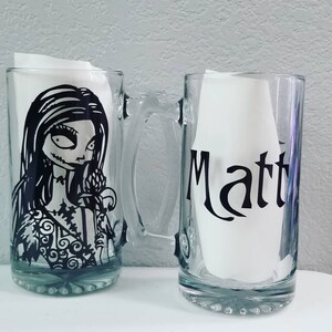 May include: Two clear glass beer mugs. One mug features a black and white illustration of a character from The Nightmare Before Christmas. The other mug has the name "Matt" in black lettering. Both mugs have handles.
