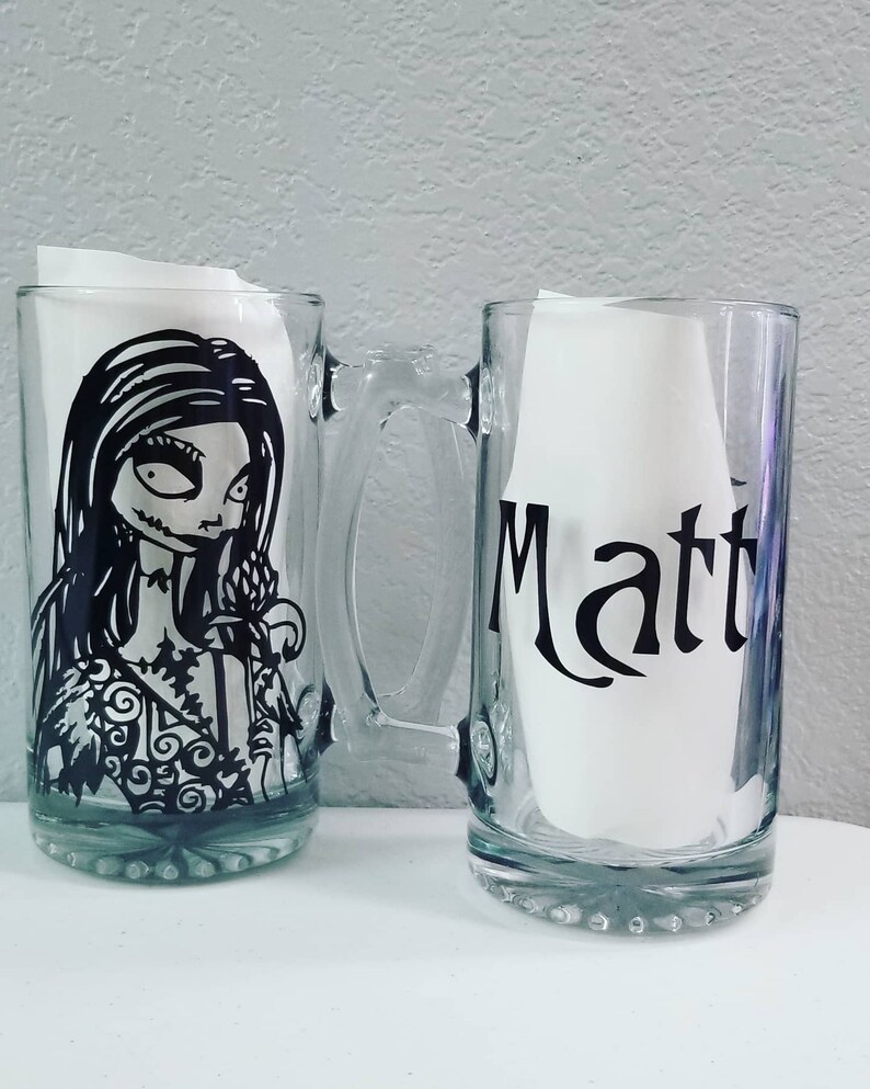 May include: Two clear glass beer mugs. One mug features a black and white illustration of a character from The Nightmare Before Christmas. The other mug has the name "Matt" in black lettering. Both mugs have handles.