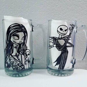 May include: Two clear glass beer mugs with black and white character designs. One mug features a woman with long hair and a floral dress, and the other features a skeleton in a pinstripe suit.