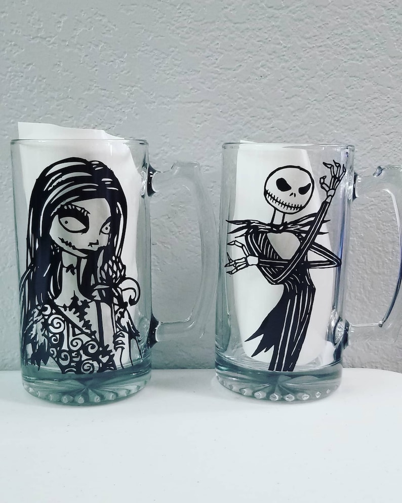 May include: Two clear glass beer mugs with black and white character designs. One mug features a woman with long hair and a floral dress, and the other features a skeleton in a pinstripe suit.
