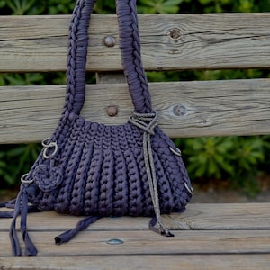 May include: A handmade, dark purple handbag with a braided handle and a textured, ribbed body. The bag features silver-toned metal rings and a decorative silver chain. The bag is sitting on a wooden bench.
