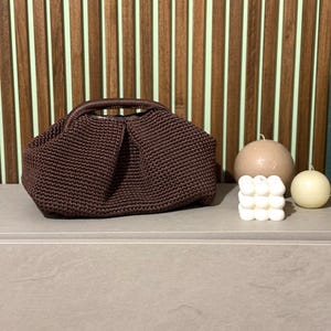 Könnte beinhalten: Eine dunkelbraune, gehäkelte Handtasche mit runder Form und Henkel. Die Tasche steht auf einer grauen Oberfläche, mit zwei Kerzen und einer dekorativen Würfelkerze in der Nähe. Der Hintergrund zeigt vertikale Holzpaneele.