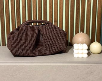 Könnte beinhalten: Eine dunkelbraune, gehäkelte Handtasche mit runder Form und Henkel. Die Tasche steht auf einer grauen Oberfläche, mit zwei Kerzen und einer dekorativen Würfelkerze in der Nähe. Der Hintergrund zeigt vertikale Holzpaneele.