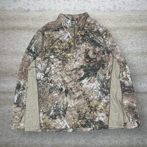Puede incluir: Una camisa de camuflaje de manga larga con cuello con cremallera de un cuarto. La camisa presenta un patrón de marrón, verde y beige, con paneles beige lisos en los laterales. La camisa está diseñada para actividades al aire libre.