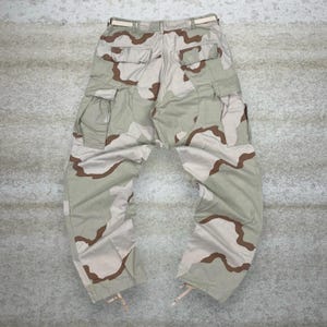 May include: Desert camouflage cargo pants with multiple pockets and a beige belt. The pants feature a tan and brown pattern, with drawstrings at the ankles. The pants are made of a durable material.