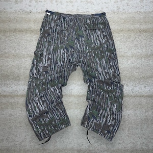 May include: Camouflage cargo pants in shades of green, gray, and brown. The pants feature a leaf and tree bark pattern, multiple pockets, and drawstrings at the ankles. The pants are laid flat on a light gray surface.