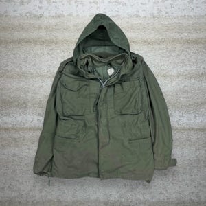 May include: Olive green military-style jacket with a hood. The jacket features multiple front pockets, a zipper closure, and a drawstring waist. The jacket is made of a durable fabric and has a vintage aesthetic.