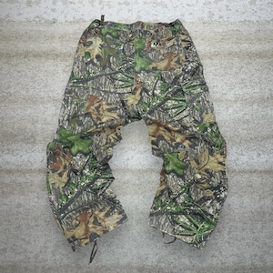 May include: Camouflage cargo pants in shades of green, brown, and tan. The pants feature multiple pockets and a drawstring at the ankles. The Mossy Oak logo is visible on the back pocket. The pants are made of a durable fabric.