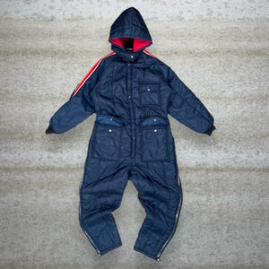 May include: A navy blue quilted snowsuit with a hood and red accents. The one-piece suit features a zip-up front, pockets, and red stripes on the sleeves. The hood has a red lining. The suit has zippers at the ankles.