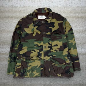 Puede incluir: Chaqueta de camuflaje en tonos verdes, marrones y beige. La chaqueta tiene cuello, cierre de botones y varios bolsillos. Se ve una etiqueta en el cuello.