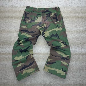 May include: Camouflage pants in shades of green, brown, and black. The pants have two back pockets and a button and zipper closure. The fabric has a military-style camouflage pattern.