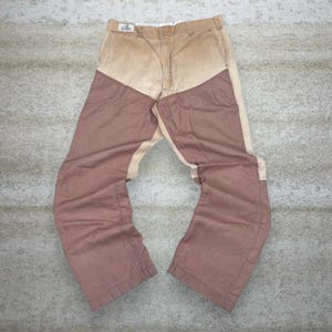May include: Tan and mauve-colored pants with a unique color-block design. The pants feature a light tan upper section and mauve panels on the legs. The pants are made of a durable fabric and have a classic, casual style.