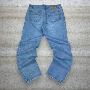 May include: Light blue denim jeans with a classic five-pocket design. The jeans have a straight leg cut and a faded wash. The back features two pockets and a leather patch. The jeans are laid flat on a neutral surface.