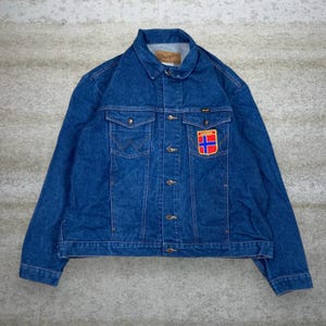 May include: A blue denim jacket with a button-up front, two chest pockets, and a collar. The jacket features a patch with a red, blue, and white design. The Wrangler logo is visible on the jacket.