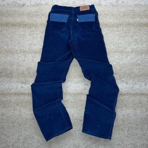 May include: Dark blue corduroy trousers with two-tone back pockets. The trousers have a classic five-pocket design and a Levi's brand patch on the back. The trousers are laid flat on a neutral surface.