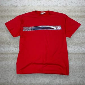 May include: A red t-shirt with a horizontal graphic across the chest. The graphic is a silver and black design with the word "SCH" visible. The shirt is made of a soft material and has short sleeves.
