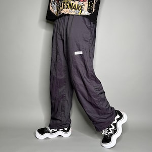 Könnte beinhalten: Dunkelgraue Nylon-Trainingshose mit kleinem weißem Logo-Patch. Die Hose hat eine lockere Passform und wird mit schwarz-weißen Turnschuhen getragen. Das Bild zeigt die Hose von der Taille abwärts.