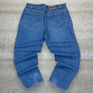 May include: Light blue denim jeans with classic five-pocket styling. The jeans feature a faded wash and a leather patch on the back waistband. The back pockets have a signature stitch design. The jeans are laid flat on a neutral surface.