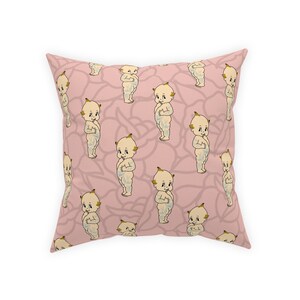 May include: Pink cushion with a repeating pattern of cartoon babies with yellow hair and blue eyes. The babies are standing with their hands clasped in front of them.