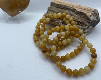 May include: A set of four yellow-brown beaded bracelets, stacked on top of a piece of driftwood. The bracelets are made of round, smooth beads.