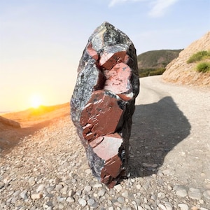May include: A large, dark gray and reddish-brown crystal with a rough, faceted surface. The crystal is sitting on a gravel road with a mountain in the background.